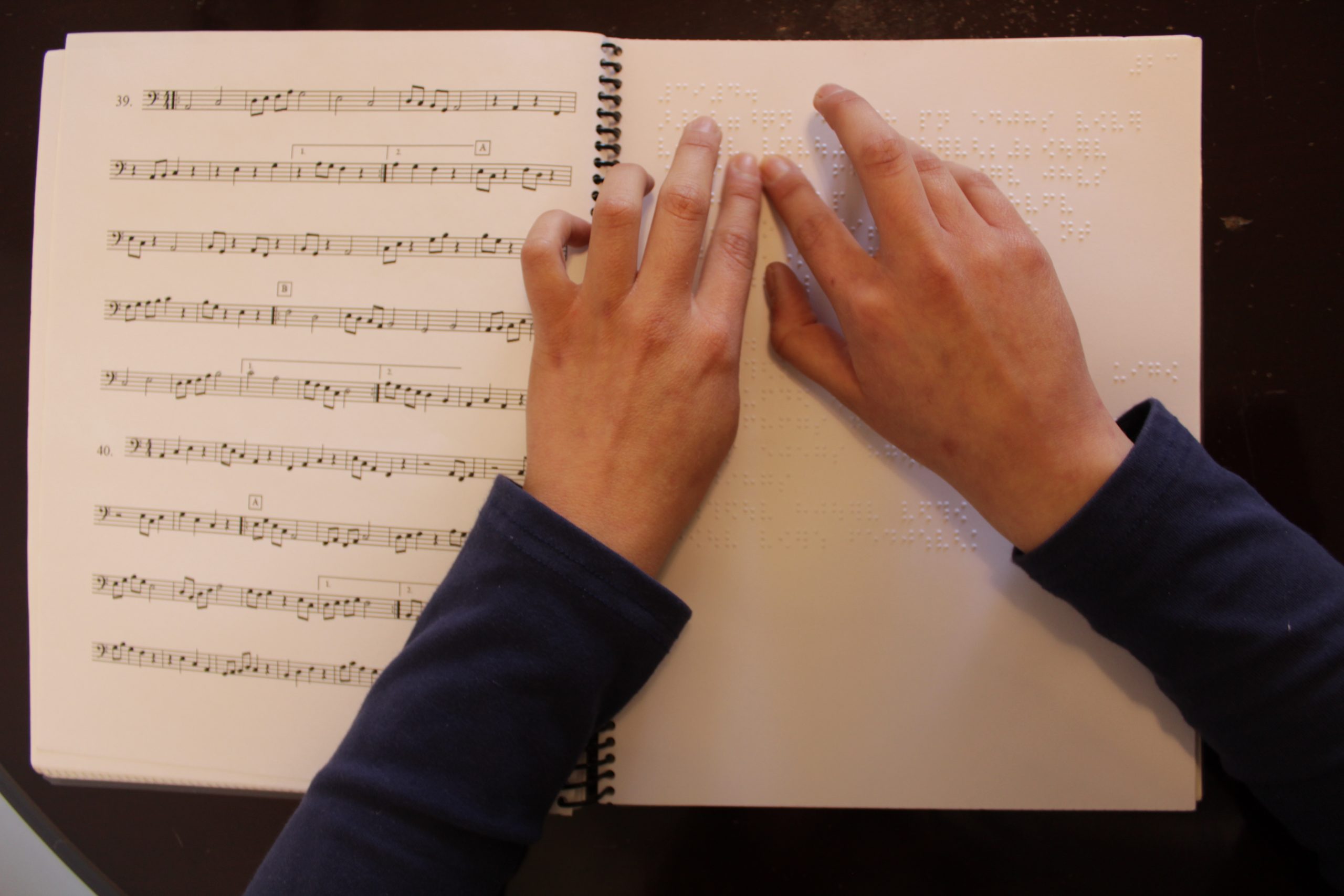 Manual de solfeo inclusivo abierto, a la izquierda partituras en tinta y a la derecha en Braille, dos manos leen el Braille.